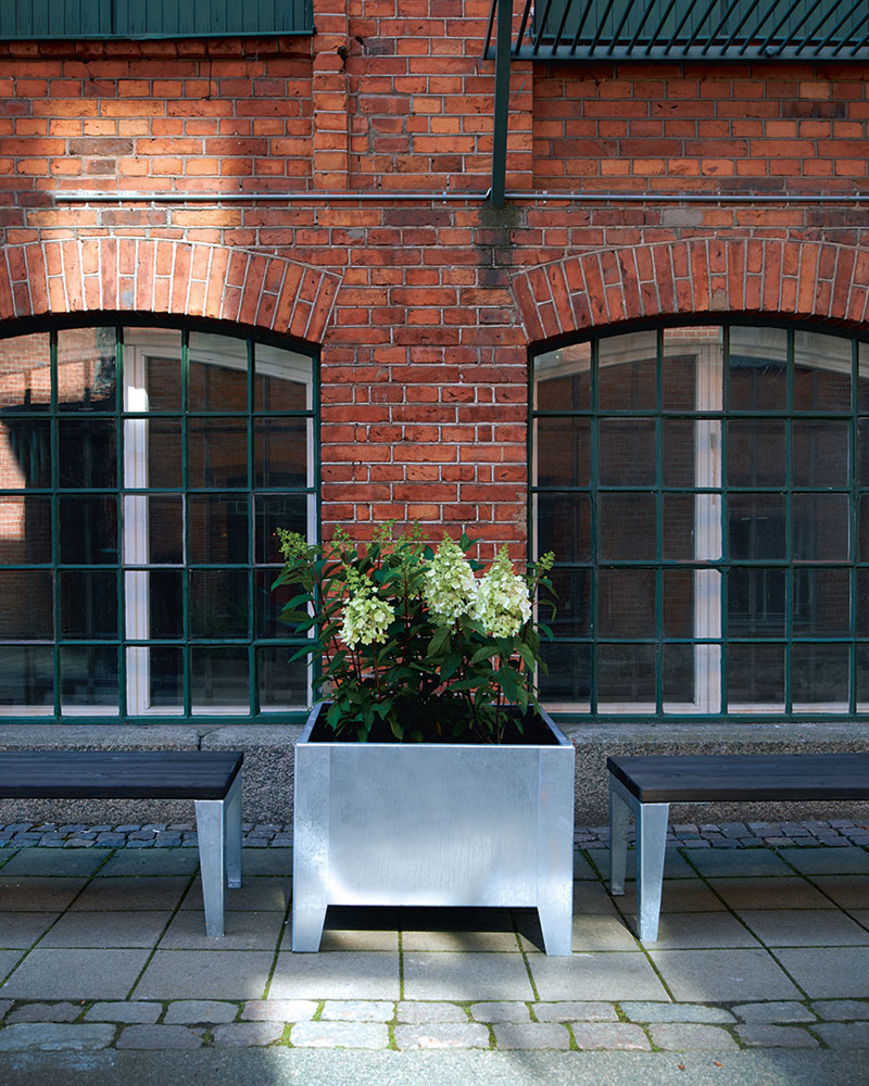 Street walkway with benches and a galvanized steel planter in the middle of two benches with plants growing within.